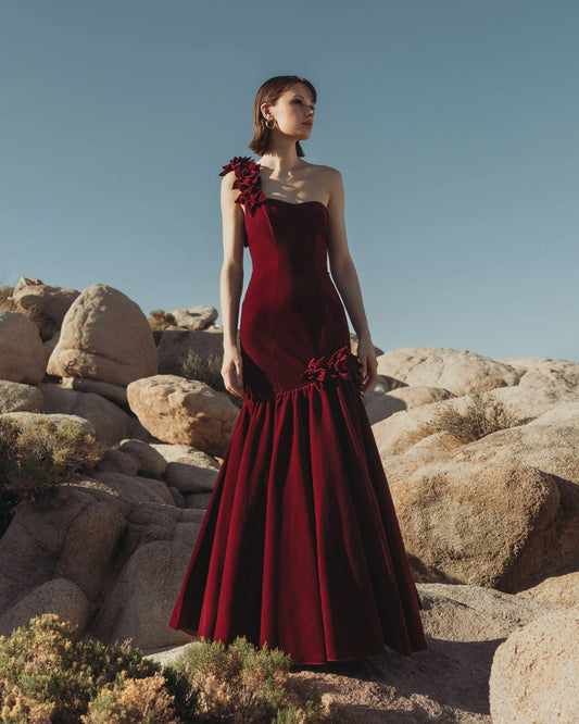 Elegant burgundy velvet mermaid gown with one-shoulder floral appliqué and dramatic flared hem, perfect for formal evening events