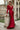 Elegant woman wearing a long red satin evening gown with a deep V-neckline and long sleeves, standing in front of a grand architectural building